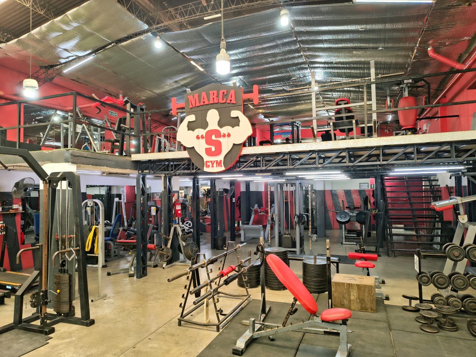 Salón principal con el cartel de Marca S Gym, peso libre y entrepiso